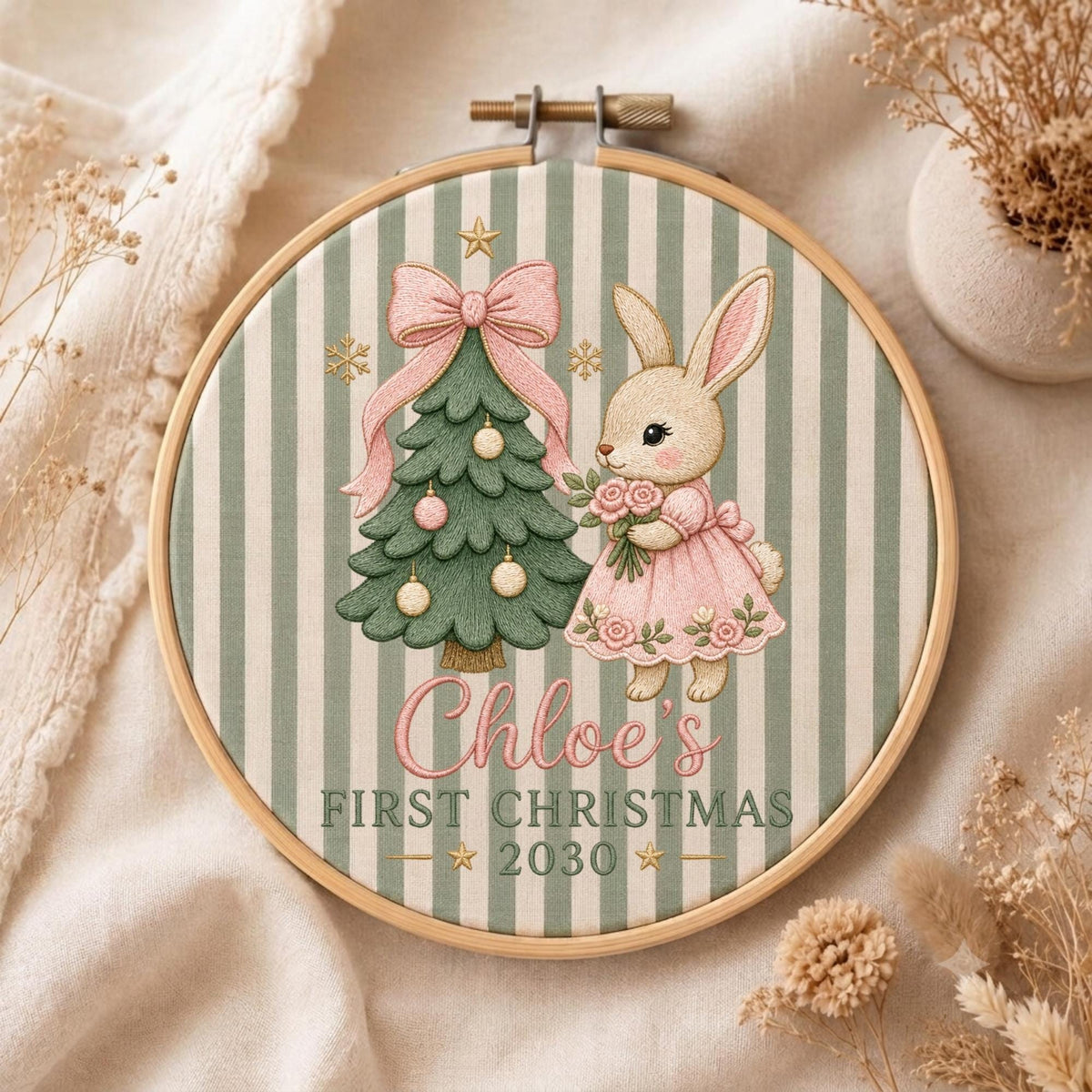 May include: A circular embroidery hoop featuring a green and white striped fabric background. The design includes a green Christmas tree with a pink bow, a rabbit in a pink dress holding flowers, and the text Chloe's FIRST CHRISTMAS 2030.