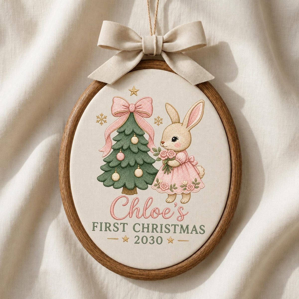 May include: An oval wooden embroidery hoop ornament featuring a cream fabric background with a printed illustration of a green Christmas tree, a pink bow, and a rabbit in a pink dress holding flowers. Text reads Chloe's FIRST CHRISTMAS 2030.
