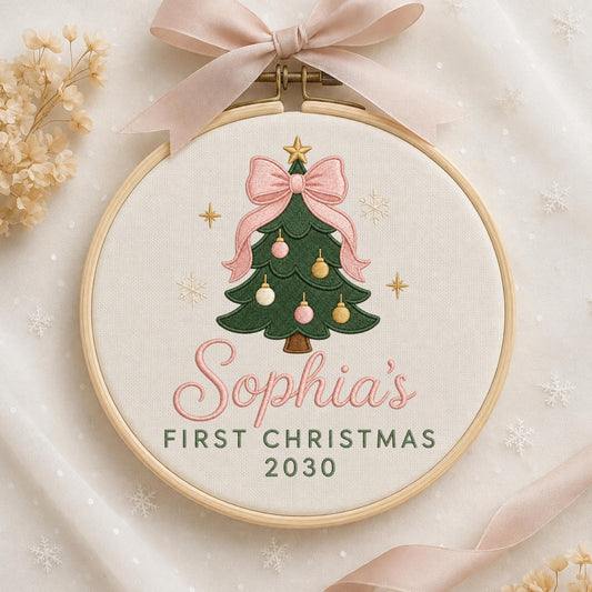 May include: A circular wooden embroidery hoop containing white fabric with a stitched green Christmas tree, pink and gold ornaments, and a pink bow. The text reads Sophia's FIRST CHRISTMAS 2030. A light pink ribbon is tied to the top of the hoop.