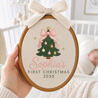 May include: An oval wooden embroidery hoop containing beige fabric with an embroidered green Christmas tree, pink bow, and ornaments. The text reads "Sophia's FIRST CHRISTMAS 2030" in pink and green. A cream-colored fabric bow is tied at the top.