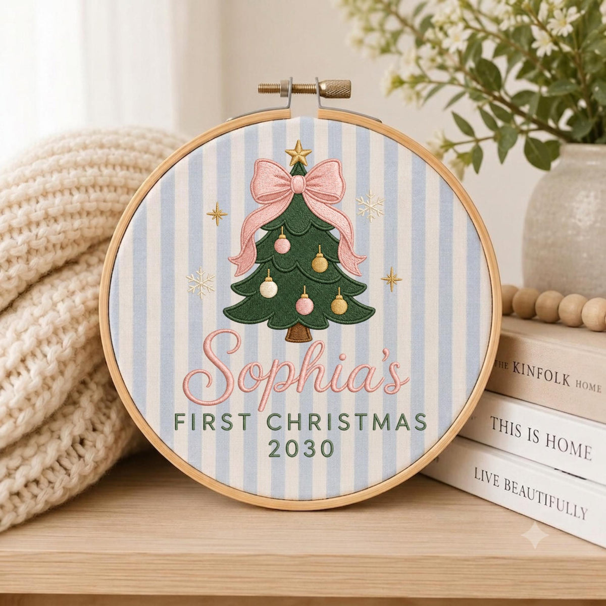 May include: A circular wooden embroidery hoop containing fabric with light blue and white vertical stripes. The center features an embroidered green Christmas tree with a pink bow, ornaments, and the text Sophia's FIRST CHRISTMAS 2030.