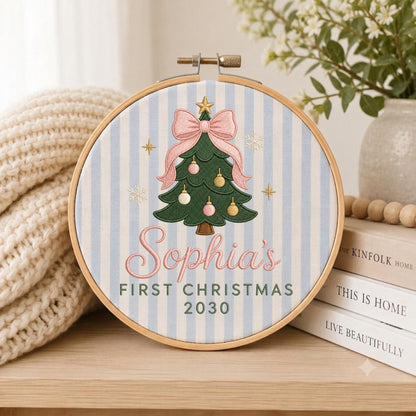 May include: A circular wooden embroidery hoop containing fabric with light blue and white vertical stripes. The center features an embroidered green Christmas tree with a pink bow, ornaments, and the text Sophia's FIRST CHRISTMAS 2030.
