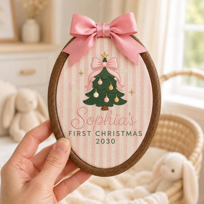 May include: An oval wooden embroidery hoop containing fabric with a pink and white striped pattern. A green Christmas tree with pink and gold ornaments is embroidered in the center, topped with a pink bow. Text reads Sophia's FIRST CHRISTMAS 2030.