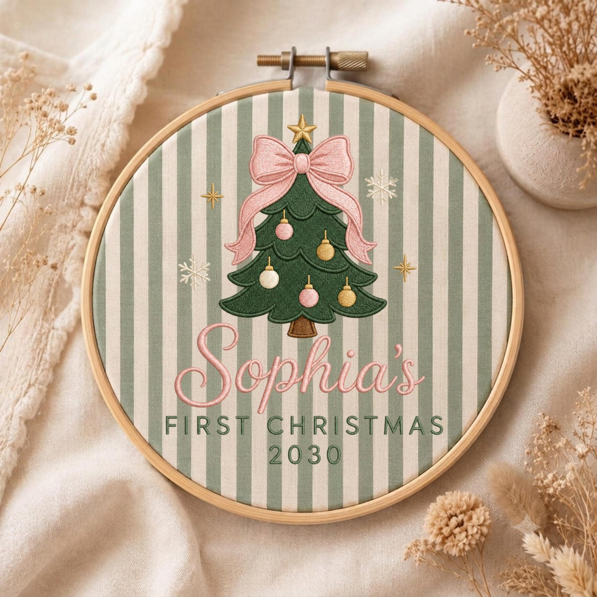 May include: A round wooden embroidery hoop containing fabric with a sage green and white vertical stripe pattern. The center features an embroidered green Christmas tree with a pink bow, gold and pink ornaments, and the text Sophia's FIRST CHRISTMAS 2030.