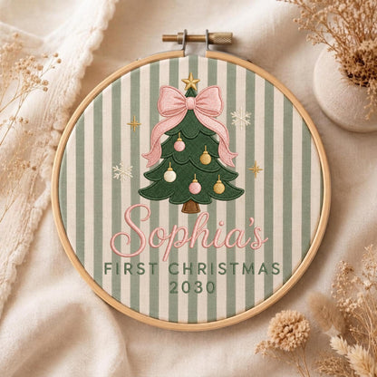 May include: A round wooden embroidery hoop containing fabric with a sage green and white vertical stripe pattern. The center features an embroidered green Christmas tree with a pink bow, gold and pink ornaments, and the text Sophia's FIRST CHRISTMAS 2030.