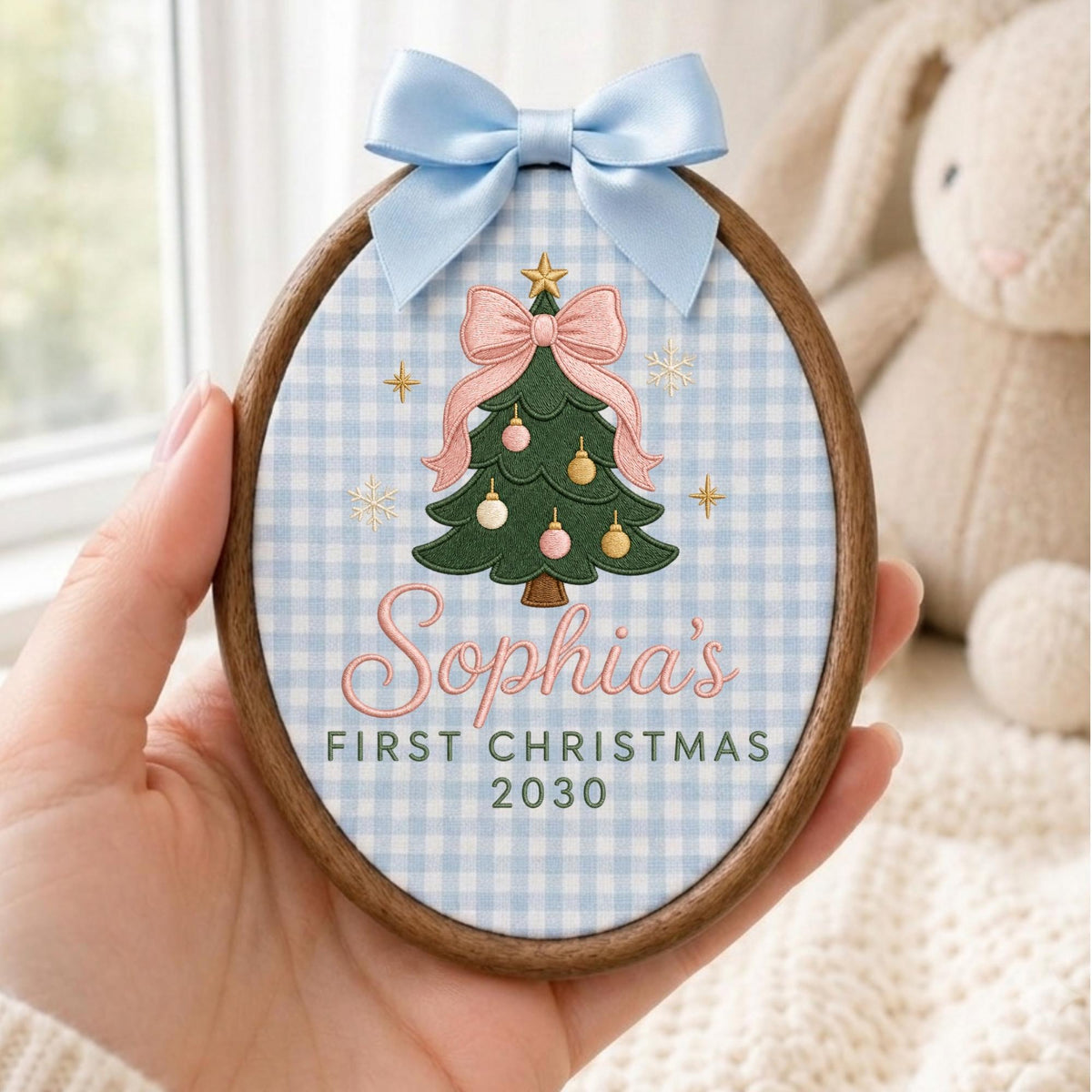 May include: An oval wooden embroidery hoop ornament featuring a light blue and white gingham fabric background. It displays an embroidered green Christmas tree with a pink bow, gold star, and ornaments, with the text Sophia's FIRST CHRISTMAS 2030.