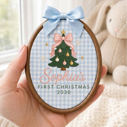 May include: An oval wooden embroidery hoop ornament featuring a light blue and white gingham fabric background. It displays an embroidered green Christmas tree with a pink bow, gold star, and ornaments, with the text Sophia's FIRST CHRISTMAS 2030.