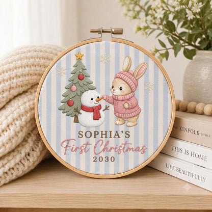 May include: A round wooden embroidery hoop containing fabric with a light blue and white vertical stripe pattern. The design features a green Christmas tree, a white snowman with a red scarf, and a brown rabbit wearing a pink sweater and hat.