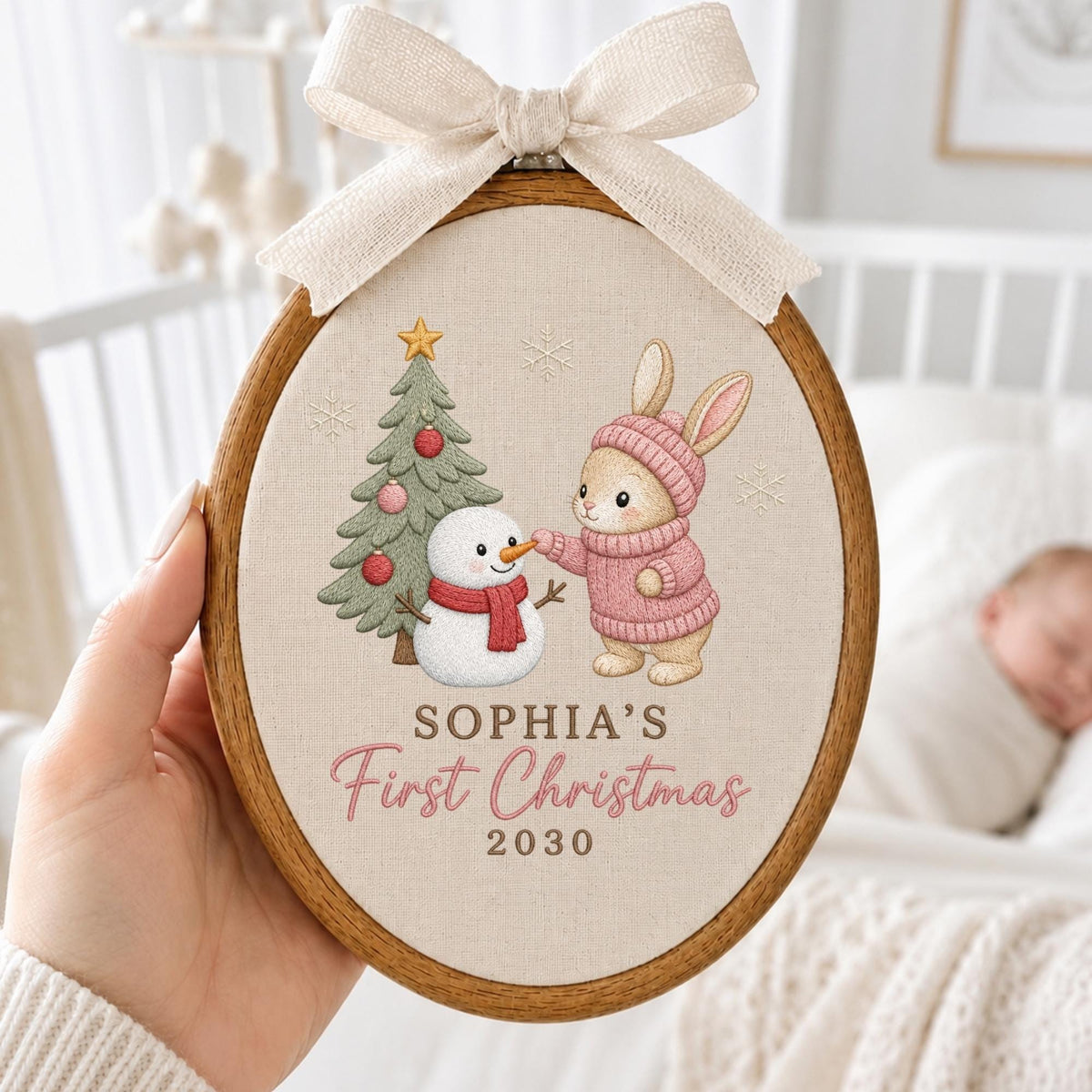 May include: An oval wooden embroidery hoop containing fabric with a printed illustration of a rabbit in a pink sweater and hat, a snowman, and a Christmas tree. The text reads SOPHIA'S First Christmas 2030. A cream-colored ribbon bow is attached.