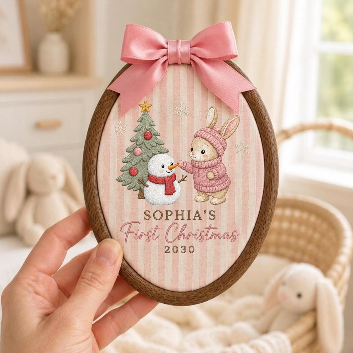 May include: An oval wooden embroidery hoop frame containing fabric with a pink and white striped pattern. The design features a cartoon rabbit in a pink sweater, a snowman, and a Christmas tree. Text reads: SOPHIA'S First Christmas 2030. A pink satin bow is attached to the top.