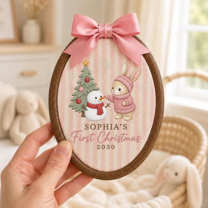 May include: An oval wooden embroidery hoop frame containing fabric with a pink and white striped pattern. The design features a cartoon rabbit in a pink sweater, a snowman, and a Christmas tree. Text reads: SOPHIA'S First Christmas 2030. A pink satin bow is attached to the top.