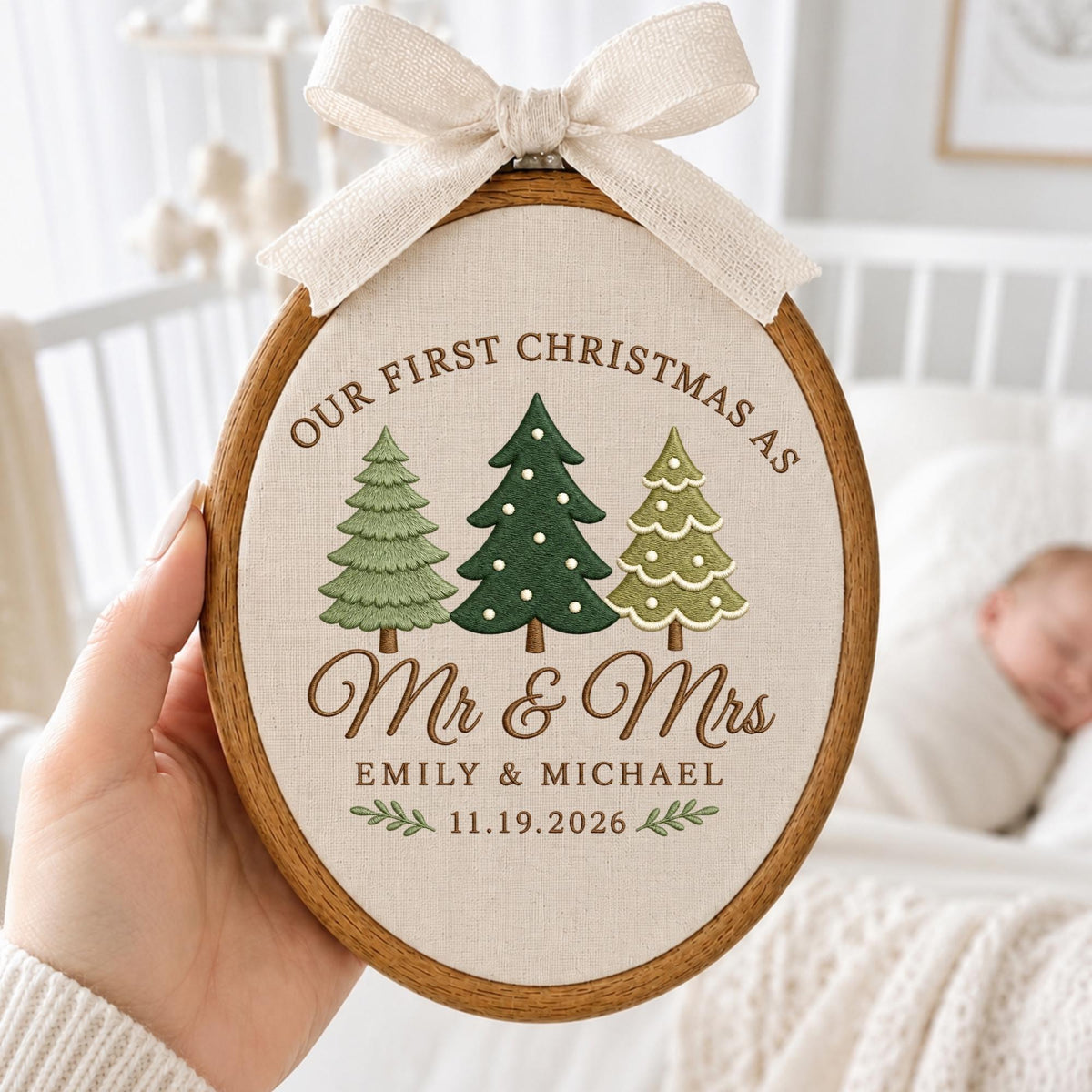 May include: An oval wooden embroidery hoop containing cream-colored fabric with embroidered text reading Our First Christmas As Mr &amp; Mrs Emily &amp; Michael 11.19.2026, three green pine trees, and a beige fabric bow attached to the top of the frame.