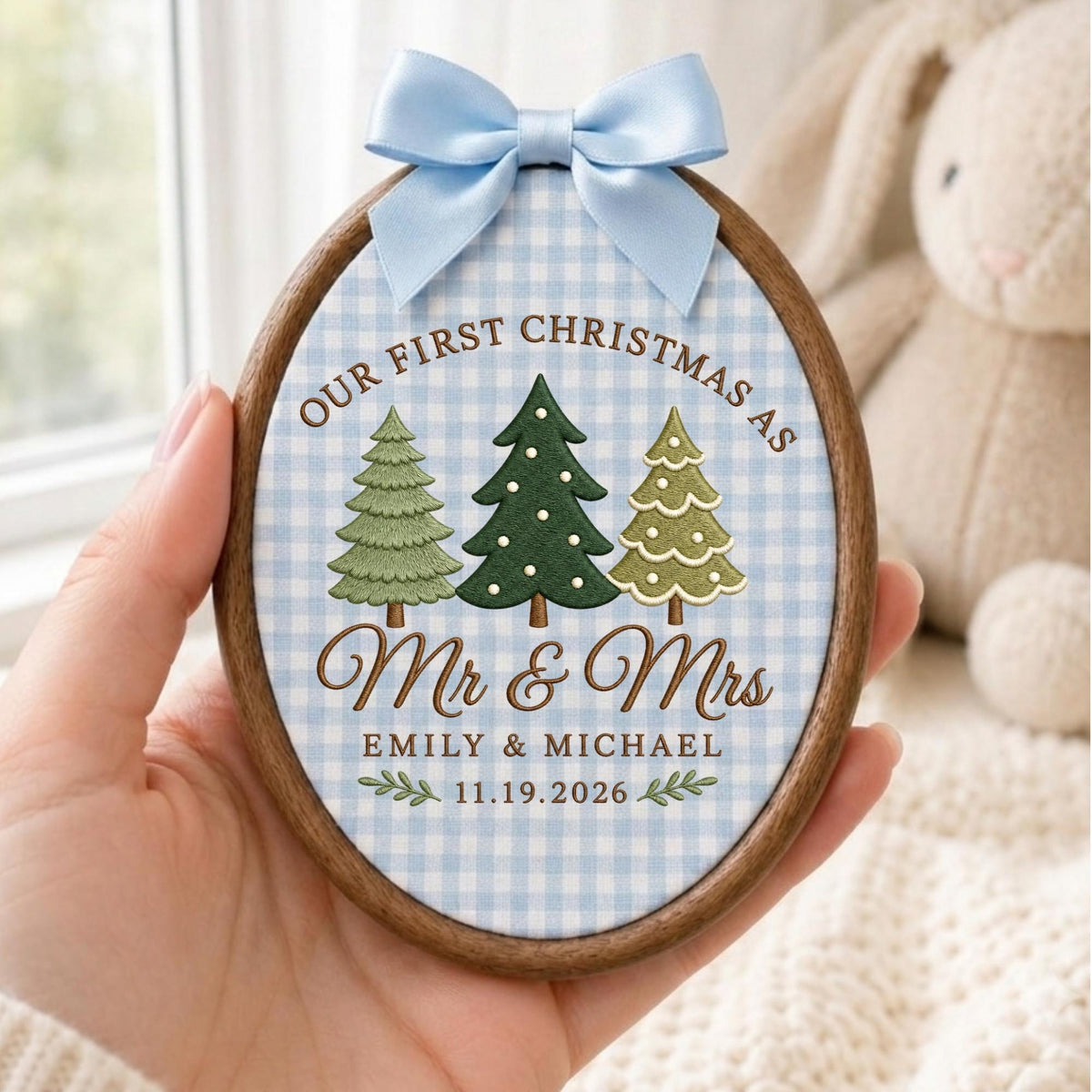 May include: An oval wooden embroidery hoop ornament featuring a light blue gingham fabric background. It displays three embroidered Christmas trees, the text "OUR FIRST CHRISTMAS AS Mr &amp; Mrs EMILY &amp; MICHAEL 11.19.2026", and a light blue satin bow.