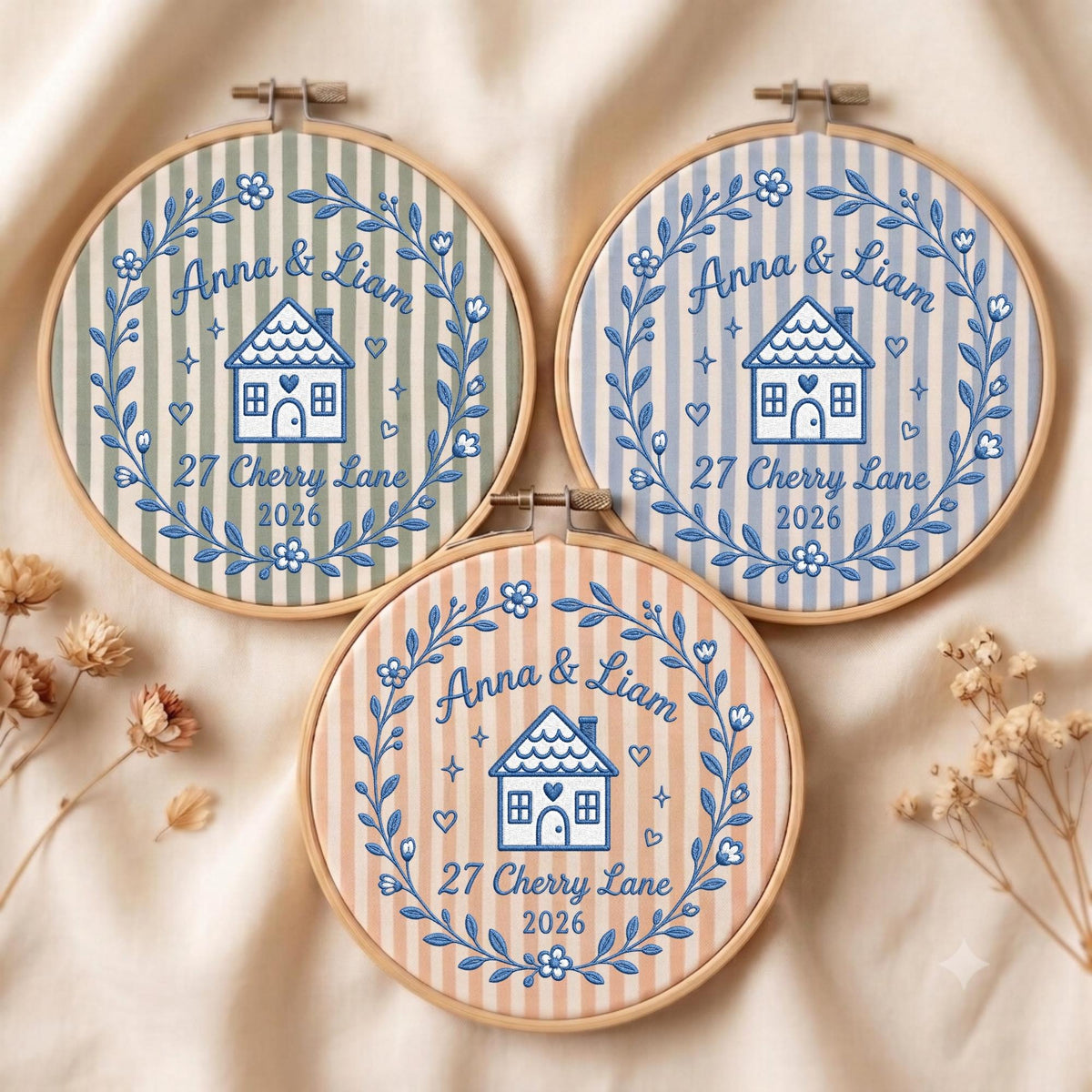 May include: Three circular wooden embroidery hoops featuring striped fabric in sage green, light blue, and peach. Each hoop displays blue embroidered text reading Anna &amp; Liam, 27 Cherry Lane, 2026, a central house icon, and a surrounding floral wreath.