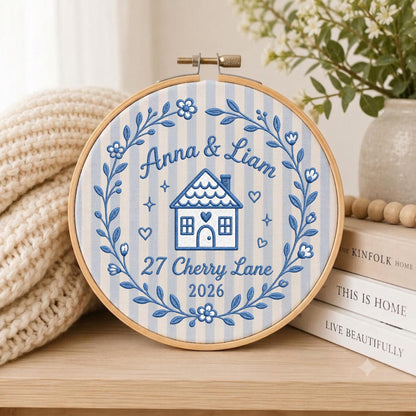May include: A round wooden embroidery hoop containing fabric with blue and white vertical stripes. The embroidery features a blue house, floral wreath, and the text Anna &amp; Liam, 27 Cherry Lane, 2026, stitched in blue thread.
