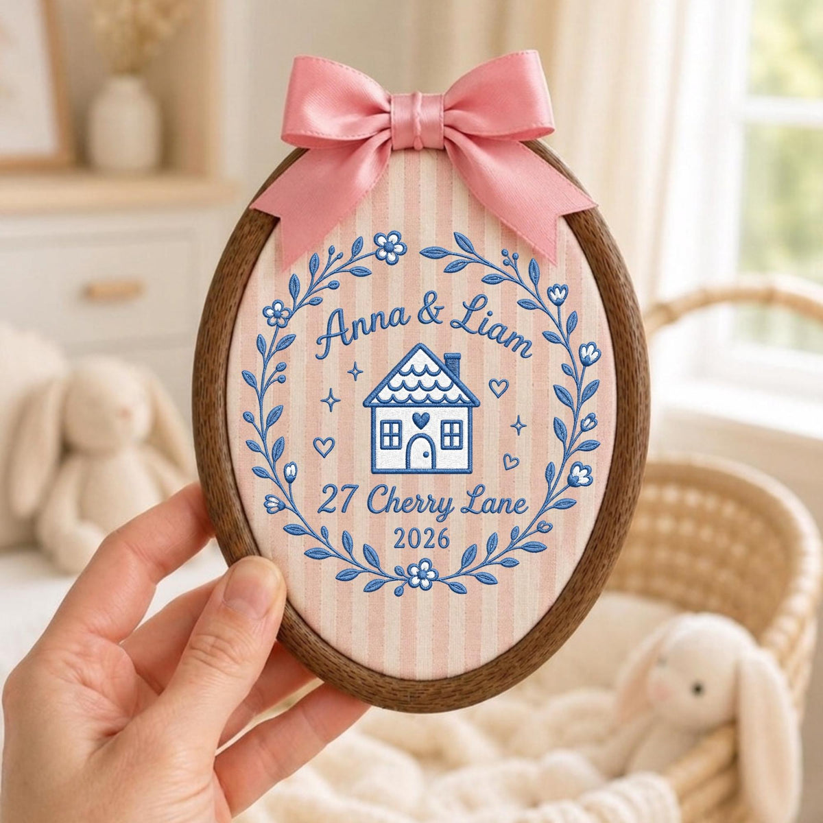 May include: An oval wooden embroidery hoop containing fabric with pink vertical stripes. The embroidery features a blue floral wreath surrounding a house icon, the names Anna &amp; Liam, the address 27 Cherry Lane, and the year 2026. A pink satin bow is attached to the top.