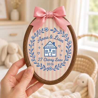 May include: An oval wooden embroidery hoop containing fabric with pink vertical stripes. The embroidery features a blue floral wreath surrounding a house icon, the names Anna &amp; Liam, the address 27 Cherry Lane, and the year 2026. A pink satin bow is attached to the top.