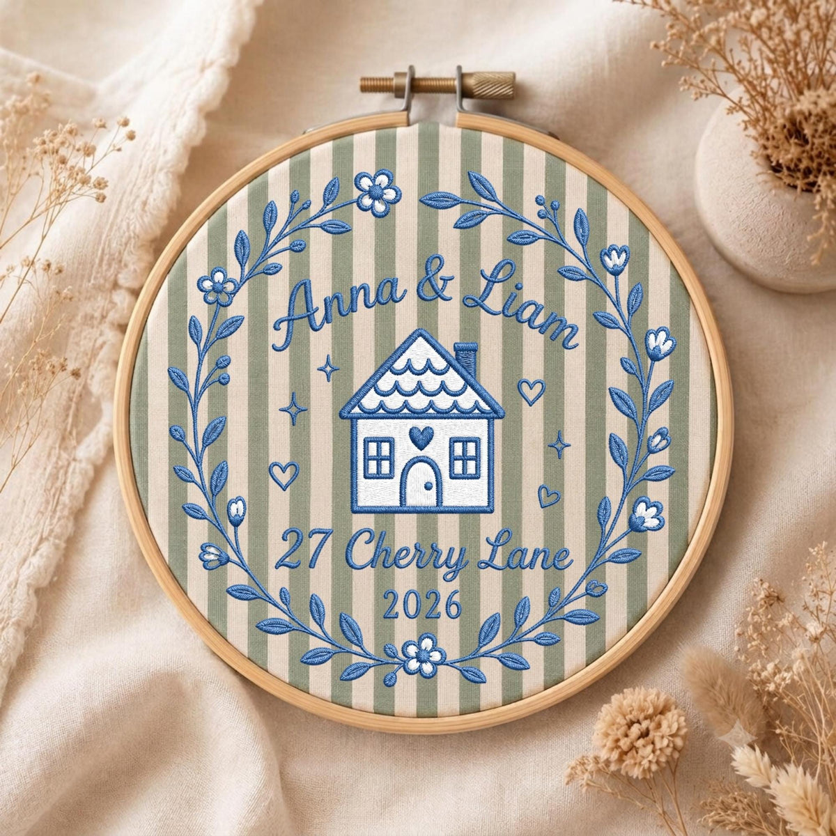 May include: A circular wooden embroidery hoop containing fabric with vertical sage green and cream stripes. Blue thread embroidery features a house, floral wreath, and the text Anna &amp; Liam, 27 Cherry Lane, 2026, accented with small hearts.