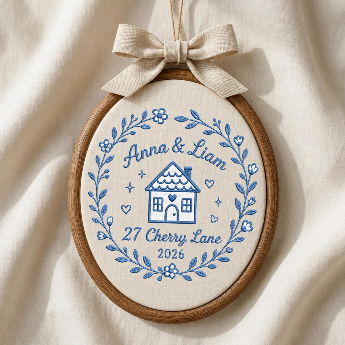 May include: An oval embroidery hoop wall hanging featuring a cream-colored fabric background with blue thread embroidery. The design includes a house, the text Anna &amp; Liam, 27 Cherry Lane, 2026, and a floral wreath. A beige fabric bow sits on top.
