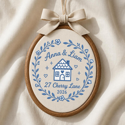 May include: An oval embroidery hoop wall hanging featuring a cream-colored fabric background with blue thread embroidery. The design includes a house, the text Anna &amp; Liam, 27 Cherry Lane, 2026, and a floral wreath. A beige fabric bow sits on top.