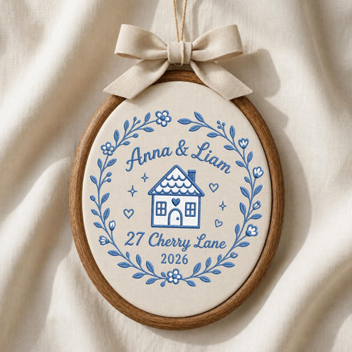 May include: An oval embroidery hoop wall hanging featuring a cream-colored fabric background with blue thread embroidery. The design includes a house, the text Anna &amp; Liam, 27 Cherry Lane, 2026, and a floral wreath. A beige fabric bow sits on top.
