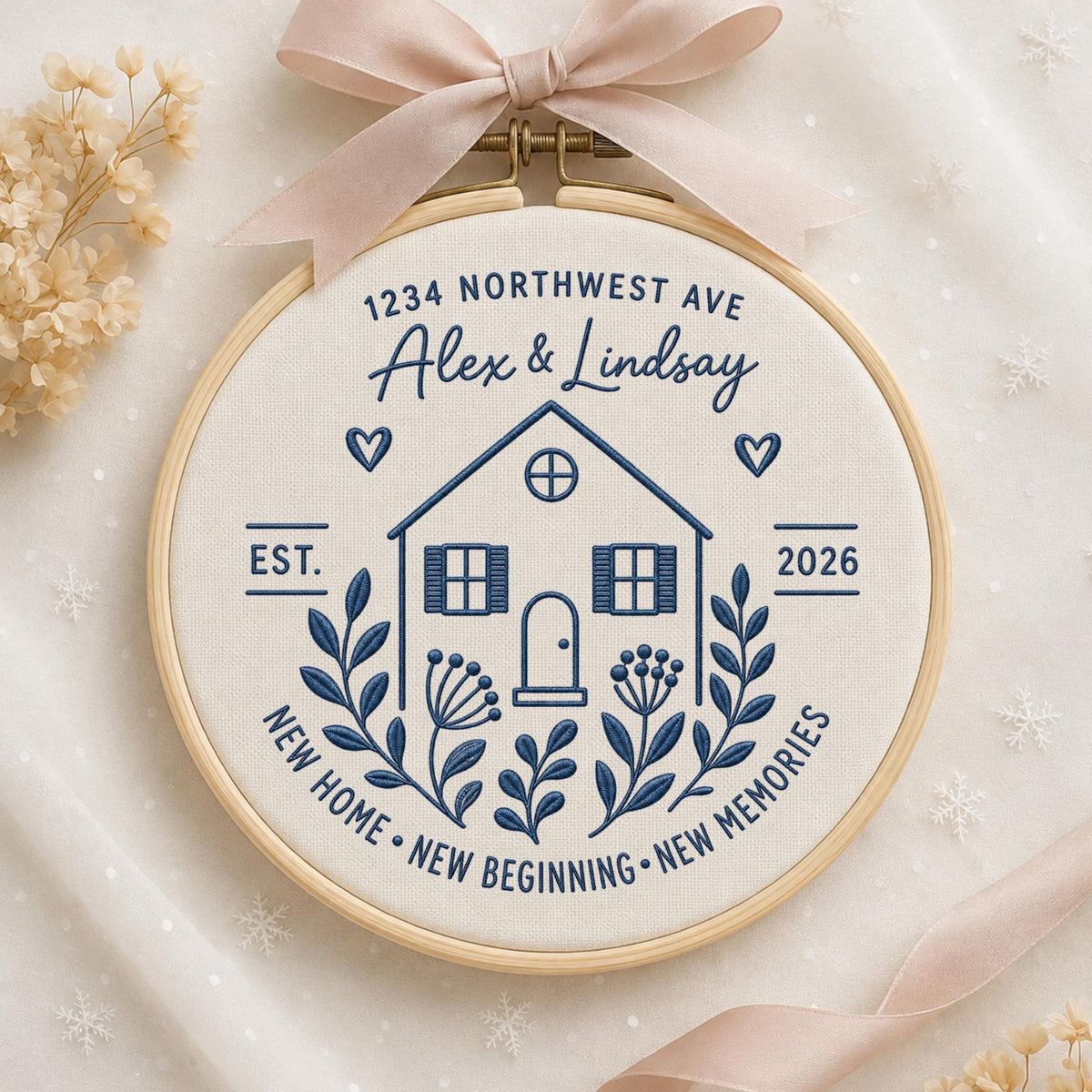 May include: A circular wooden embroidery hoop containing cream-colored fabric with dark blue stitched text and a house illustration. The text reads 1234 NORTHWEST AVE, Alex &amp; Lindsay, EST. 2026, and NEW HOME NEW BEGINNING NEW MEMORIES. A light pink ribbon is tied at the top.