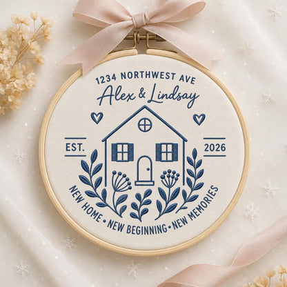 May include: A circular wooden embroidery hoop containing cream-colored fabric with dark blue stitched text and a house illustration. The text reads 1234 NORTHWEST AVE, Alex &amp; Lindsay, EST. 2026, and NEW HOME NEW BEGINNING NEW MEMORIES. A light pink ribbon is tied at the top.