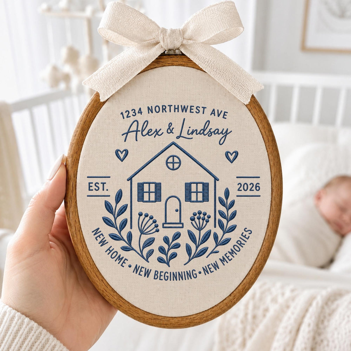 May include: An oval wooden embroidery hoop containing cream fabric with blue stitched text and graphics. The design features a house, floral sprigs, hearts, and the text 1234 NORTHWEST AVE, Alex &amp; Lindsay, EST. 2026, NEW HOME, NEW BEGINNING, NEW MEMORIES. A beige ribbon bow is attached to the top.