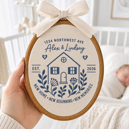 May include: An oval wooden embroidery hoop containing cream fabric with blue stitched text and graphics. The design features a house, floral sprigs, hearts, and the text 1234 NORTHWEST AVE, Alex &amp; Lindsay, EST. 2026, NEW HOME, NEW BEGINNING, NEW MEMORIES. A beige ribbon bow is attached to the top.