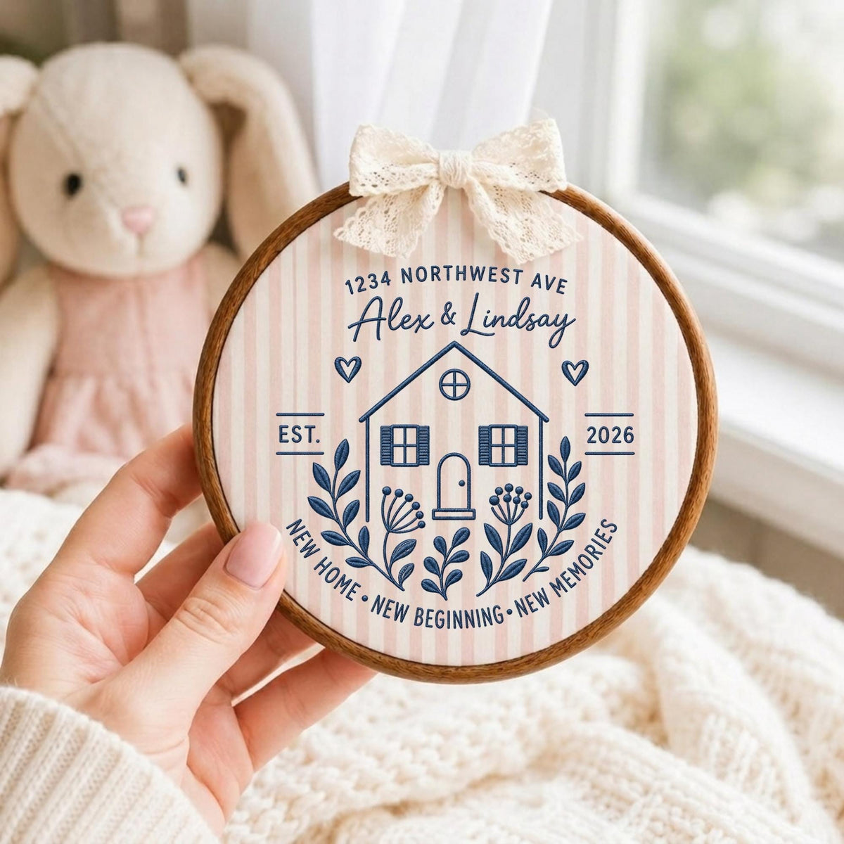 May include: A circular wooden embroidery hoop containing fabric with vertical light pink stripes. The design features a dark blue house illustration, floral accents, and text reading 1234 NORTHWEST AVE, Alex &amp; Lindsay, EST. 2026, NEW HOME, NEW BEGINNING, NEW MEMORIES. A cream lace bow is attached to the top.