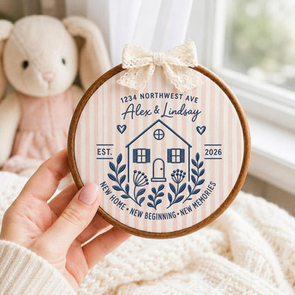 May include: A circular wooden embroidery hoop containing fabric with vertical light pink stripes. The design features a dark blue house illustration, floral accents, and text reading 1234 NORTHWEST AVE, Alex &amp; Lindsay, EST. 2026, NEW HOME, NEW BEGINNING, NEW MEMORIES. A cream lace bow is attached to the top.