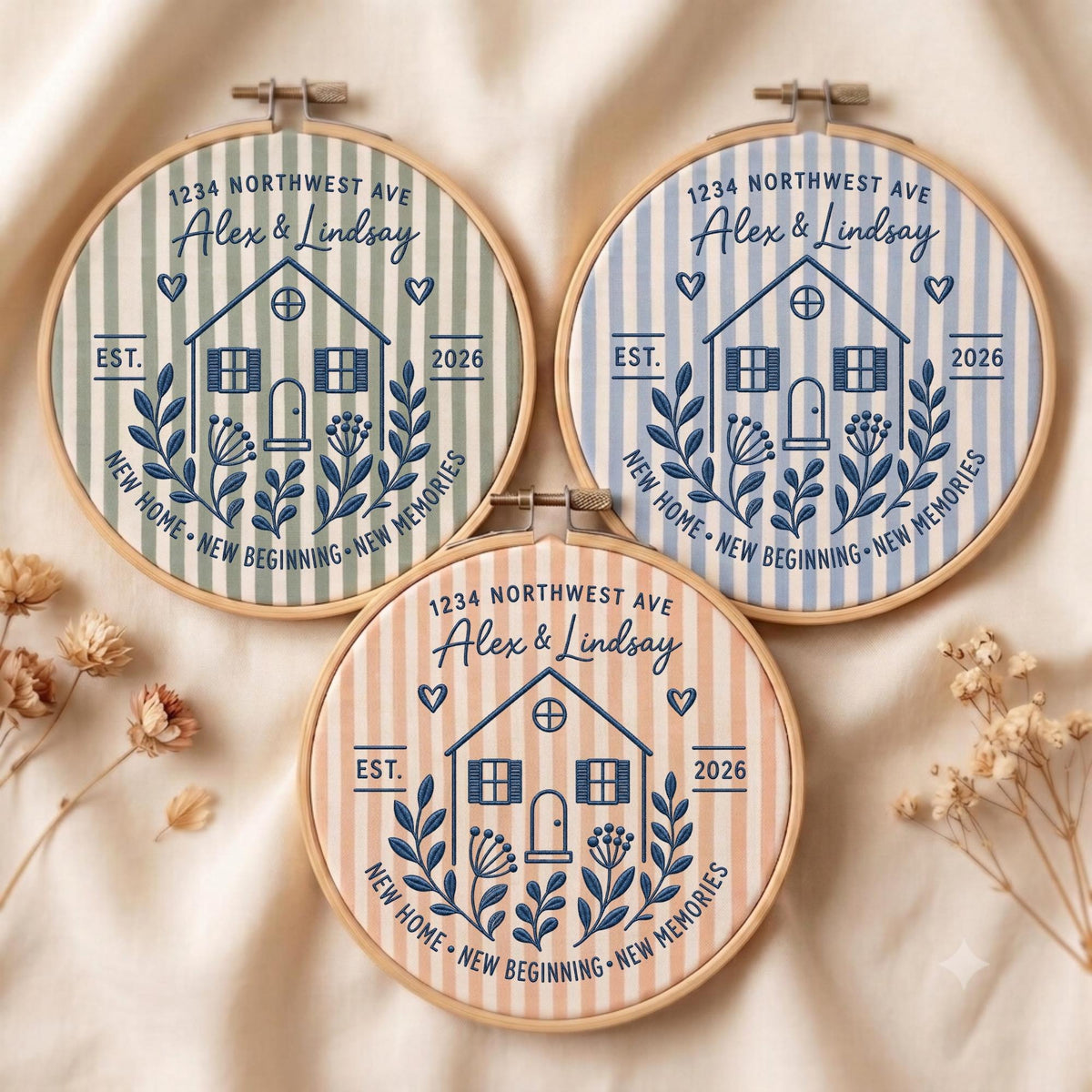 May include: Three circular embroidery hoops featuring striped fabric in sage green, light blue, and peach. Each hoop displays dark blue embroidery of a house, the text 1234 NORTHWEST AVE, Alex &amp; Lindsay, EST. 2026, and NEW HOME NEW BEGINNING NEW MEMORIES.
