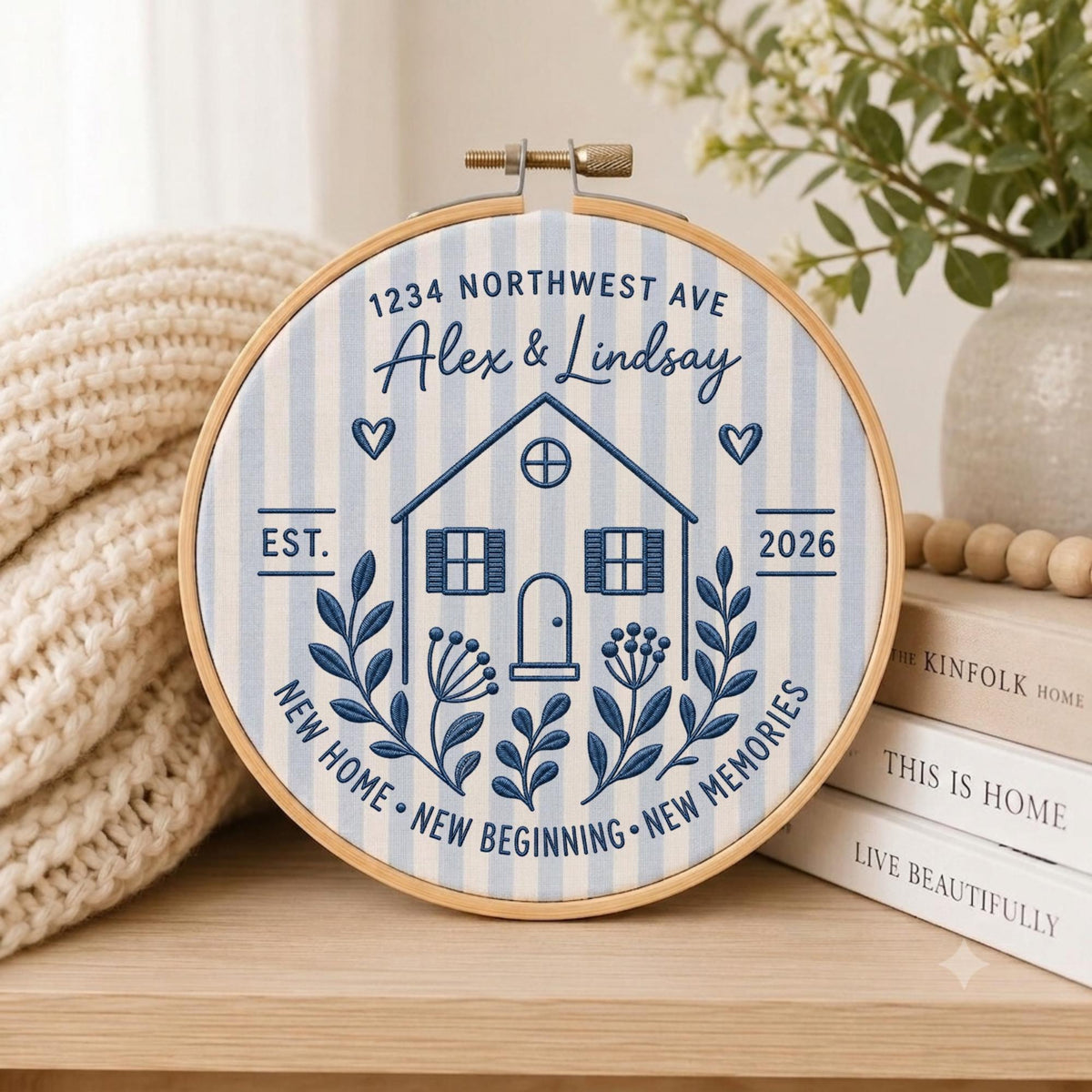 May include: A circular embroidery hoop containing fabric with light blue and white vertical stripes. The design features a dark blue embroidered house, the text 1234 NORTHWEST AVE, Alex &amp; Lindsay, EST. 2026, and NEW HOME NEW BEGINNING NEW MEMORIES.