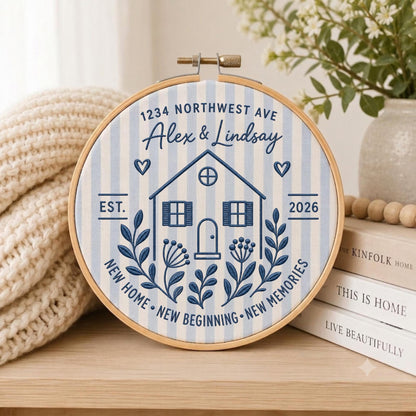 May include: A circular embroidery hoop containing fabric with light blue and white vertical stripes. The design features a dark blue embroidered house, the text 1234 NORTHWEST AVE, Alex &amp; Lindsay, EST. 2026, and NEW HOME NEW BEGINNING NEW MEMORIES.