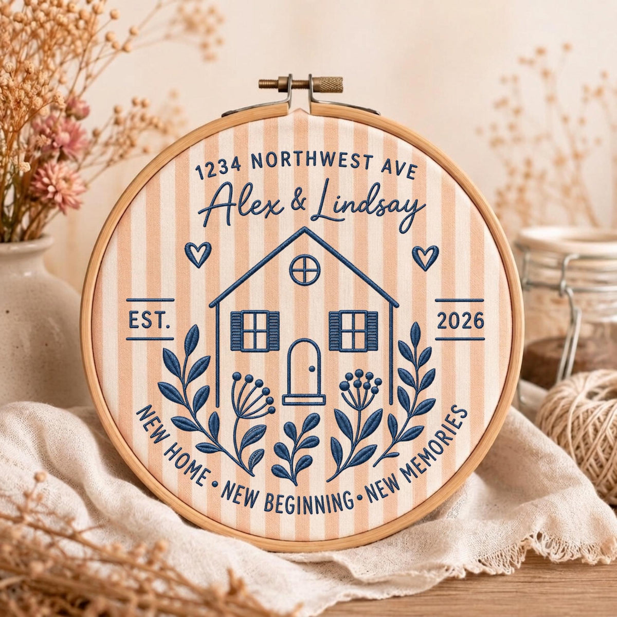 May include: A circular wooden embroidery hoop framing a light peach and white striped fabric. The embroidery features a dark blue house outline, the text 1234 NORTHWEST AVE, Alex &amp; Lindsay, EST. 2026, and NEW HOME NEW BEGINNING NEW MEMORIES.