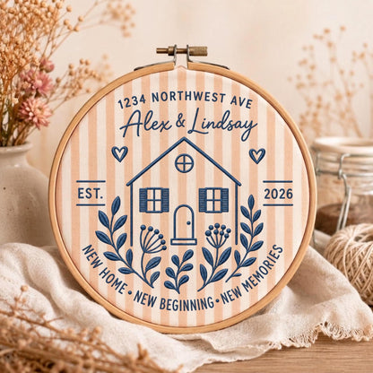 May include: A circular wooden embroidery hoop framing a light peach and white striped fabric. The embroidery features a dark blue house outline, the text 1234 NORTHWEST AVE, Alex &amp; Lindsay, EST. 2026, and NEW HOME NEW BEGINNING NEW MEMORIES.