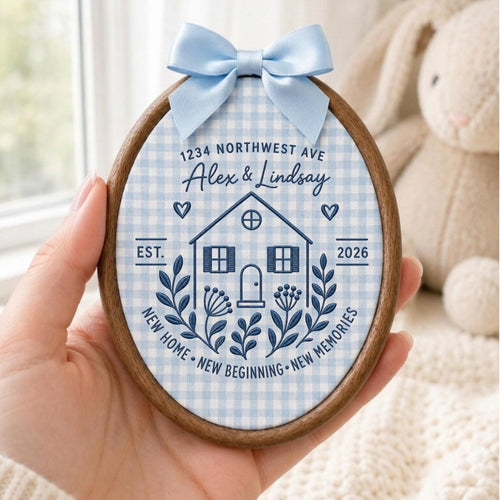 May include: An oval wooden embroidery hoop containing fabric with a light blue gingham pattern. The design features a dark blue house illustration, the text 1234 NORTHWEST AVE, Alex &amp; Lindsay, EST. 2026, and NEW HOME NEW BEGINNING NEW MEMORIES. A light blue satin bow is attached to the top.