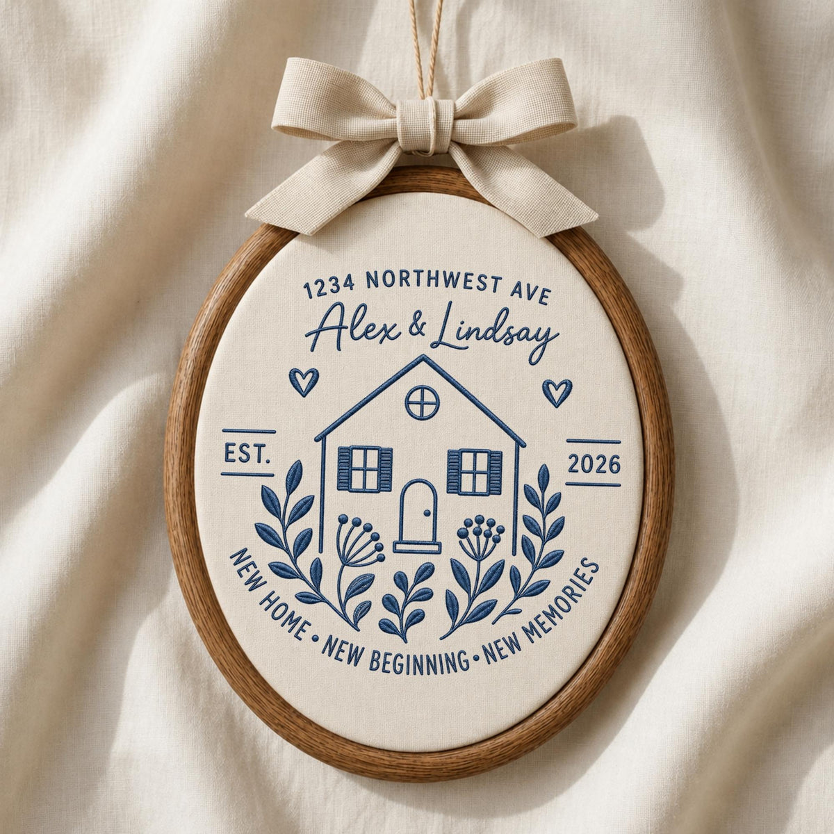 May include: An oval embroidery hoop featuring cream fabric with dark blue stitched text and graphics. The design includes a house, floral motifs, hearts, and the text 1234 NORTHWEST AVE, Alex &amp; Lindsay, EST. 2026, NEW HOME, NEW BEGINNING, NEW MEMORIES. A beige ribbon bow is attached to the top.