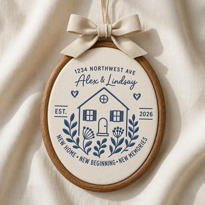 May include: An oval embroidery hoop featuring cream fabric with dark blue stitched text and graphics. The design includes a house, floral motifs, hearts, and the text 1234 NORTHWEST AVE, Alex &amp; Lindsay, EST. 2026, NEW HOME, NEW BEGINNING, NEW MEMORIES. A beige ribbon bow is attached to the top.