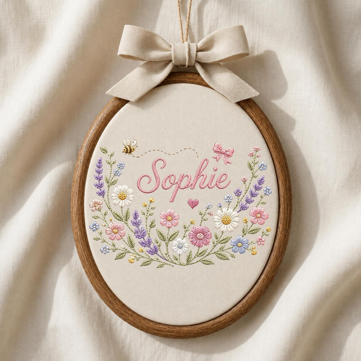 May include: An oval embroidery hoop containing cream fabric with the name Sophie stitched in pink cursive. The design features a floral wreath with daisies, lavender, and small pink flowers, accented by a small bee and a pink bow at the top.