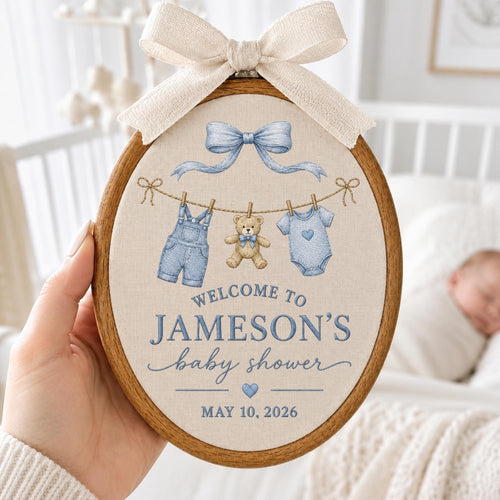 May include: An oval wooden embroidery hoop featuring a beige fabric insert with a light blue bow, a teddy bear, denim overalls, and a baby onesie hanging on a line. Text reads: WELCOME TO JAMESON'S baby shower MAY 10, 2026. A cream ribbon bow sits on top.
