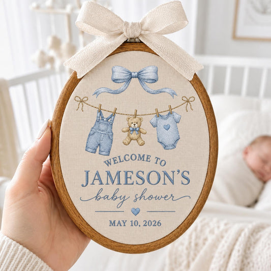 May include: An oval wooden embroidery hoop featuring a beige fabric insert with a light blue bow, a teddy bear, denim overalls, and a baby onesie hanging on a line. Text reads: WELCOME TO JAMESON'S baby shower MAY 10, 2026. A cream ribbon bow sits on top.