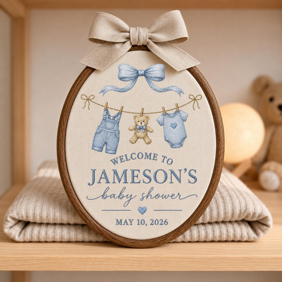 May include: An oval wooden embroidery hoop containing cream fabric with a beige bow at the top. The design features a blue bow, a clothesline with blue overalls, a teddy bear, and a blue onesie. Text reads: WELCOME TO JAMESON'S baby shower MAY 10, 2026.