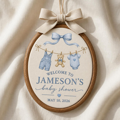 May include: An oval wooden embroidery hoop wall hanging featuring a beige fabric center with a light blue bow, a teddy bear, blue overalls, and a blue onesie. The text reads: WELCOME TO JAMESON'S baby shower MAY 10, 2026. A beige ribbon bow is at the top.