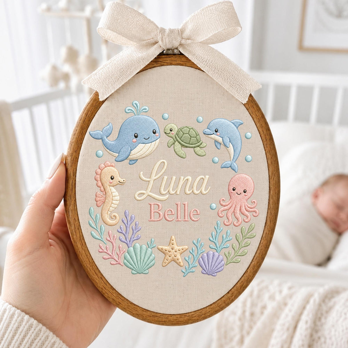 May include: An oval wooden embroidery hoop containing beige fabric with embroidered sea creatures including a blue whale, green turtle, blue dolphin, peach seahorse, and pink octopus. The name Luna Belle is stitched in the center with a beige bow.