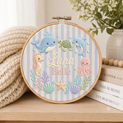 May include: A circular wooden embroidery hoop containing fabric with light blue and white vertical stripes. The fabric features embroidered sea creatures including a whale, turtle, dolphin, seahorse, octopus, starfish, and shells, with the text Luna Belle.