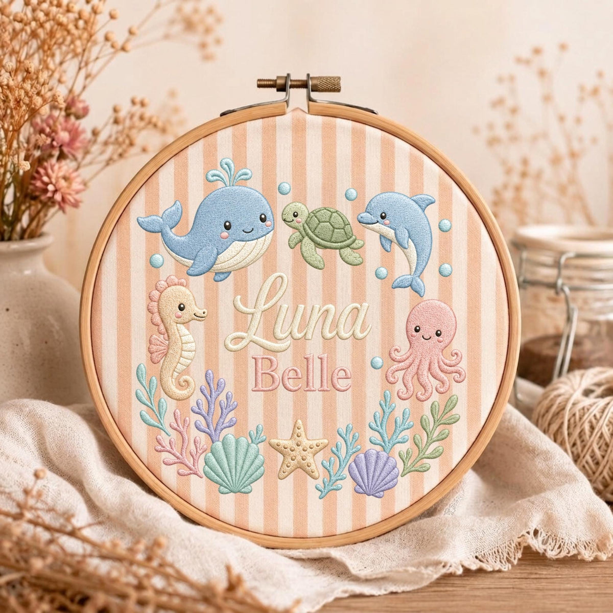 May include: A round wooden embroidery hoop containing fabric with vertical peach and white stripes. The fabric features embroidered sea creatures including a blue whale, green turtle, dolphin, seahorse, octopus, starfish, and shells with the text Luna Belle.