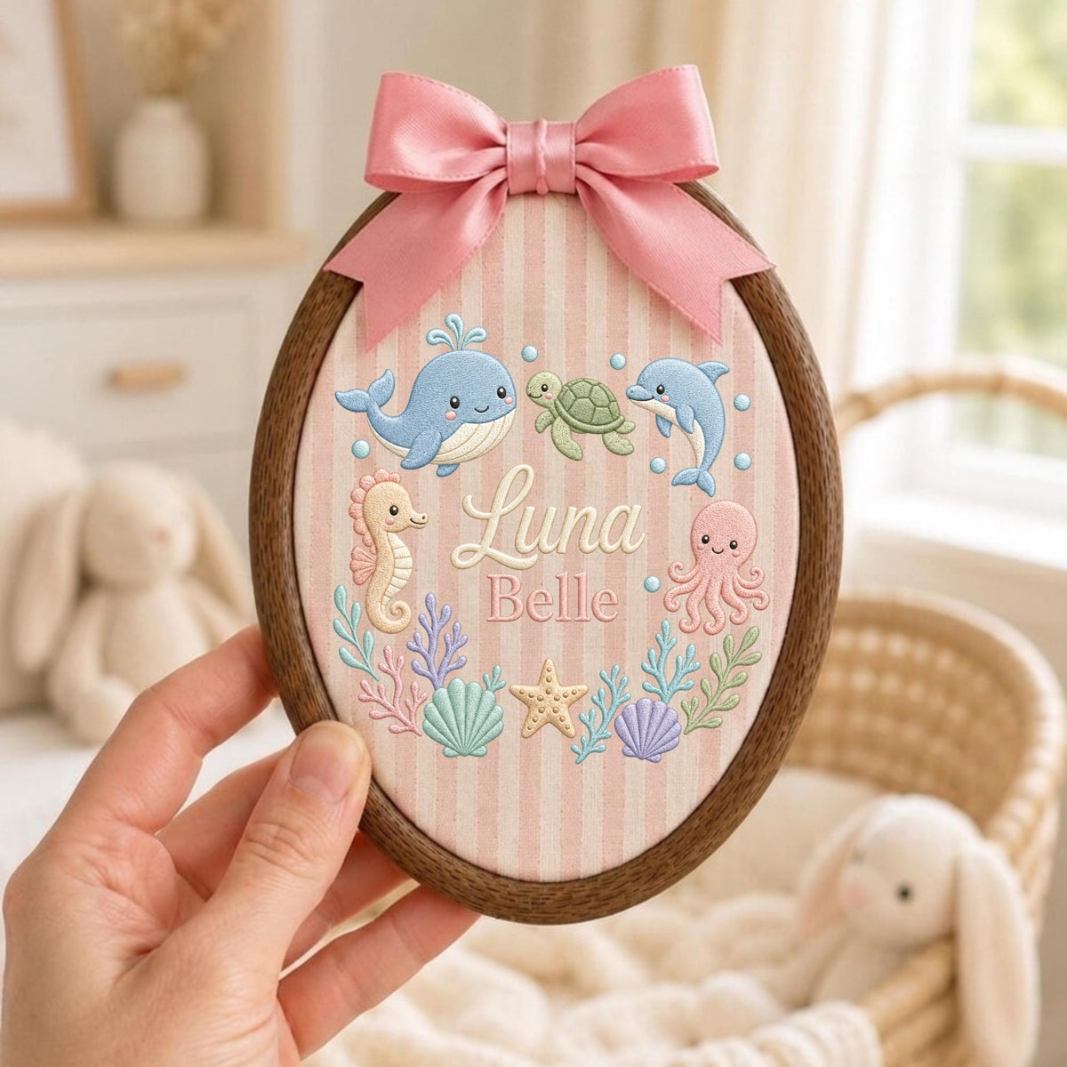 May include: An oval wooden embroidery hoop containing fabric with vertical pink stripes. The fabric features embroidered sea creatures including a whale, turtle, dolphin, seahorse, and octopus, with the name Luna Belle in the center and a pink satin bow.