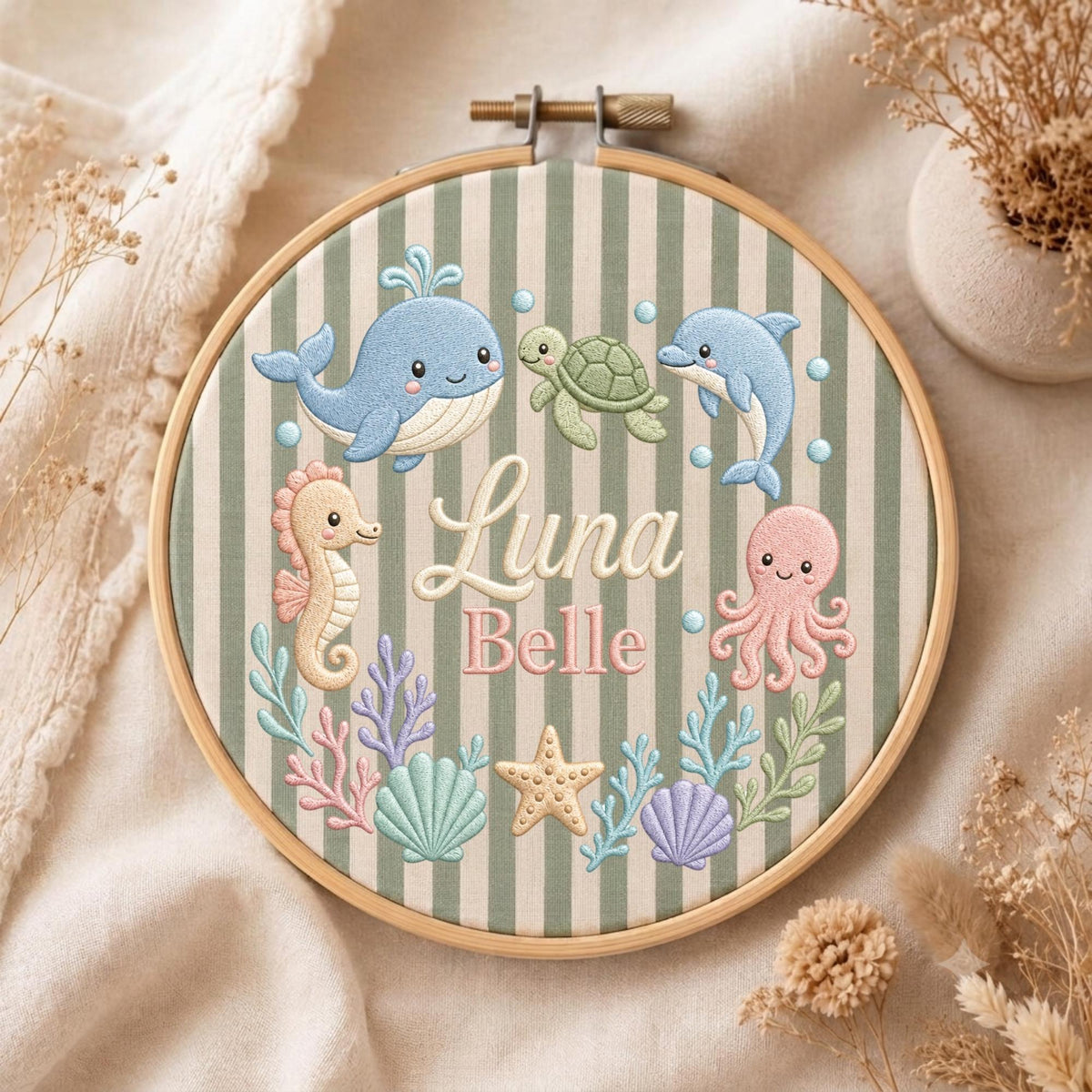 May include: A circular wooden embroidery hoop containing fabric with vertical sage green and white stripes. The embroidery features a blue whale, green turtle, blue dolphin, pink seahorse, pink octopus, starfish, shells, and coral. The text reads Luna Belle.