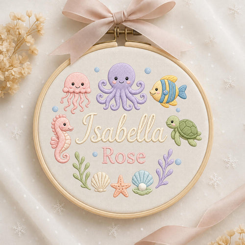 May include: A circular wooden embroidery hoop featuring a light-colored fabric background with embroidered sea creatures including a pink jellyfish, purple octopus, yellow fish, pink seahorse, and green turtle. The text Isabella Rose is centered.